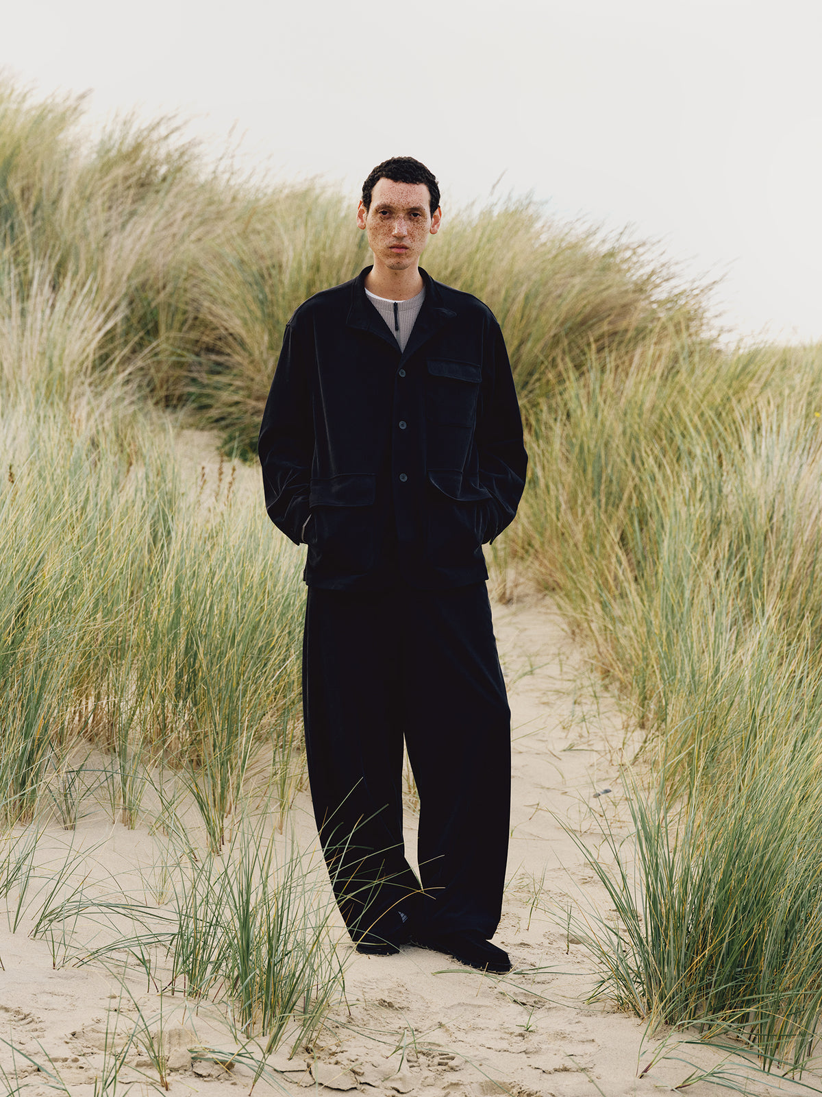 Model on a sandy path between tall beach grass, wearing a black KANEMASA PHIL jacket and matching wide-leg trousers over a Goldwin light gray shirt.