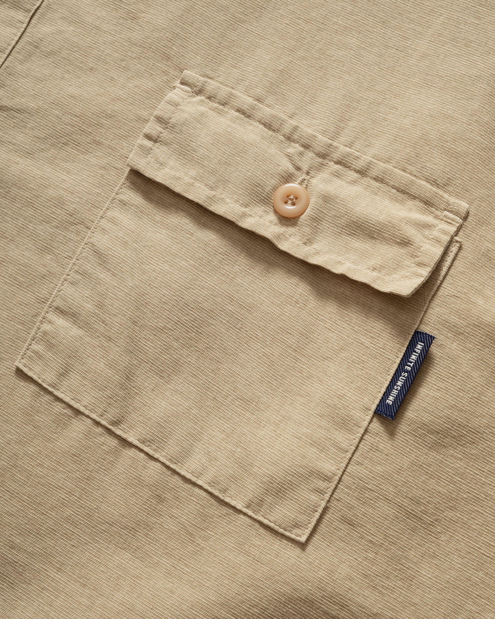 Close-up of a Pigment Henleyneck Half Shirt beige pocket with a button and a visible brand tag on a matching fabric.