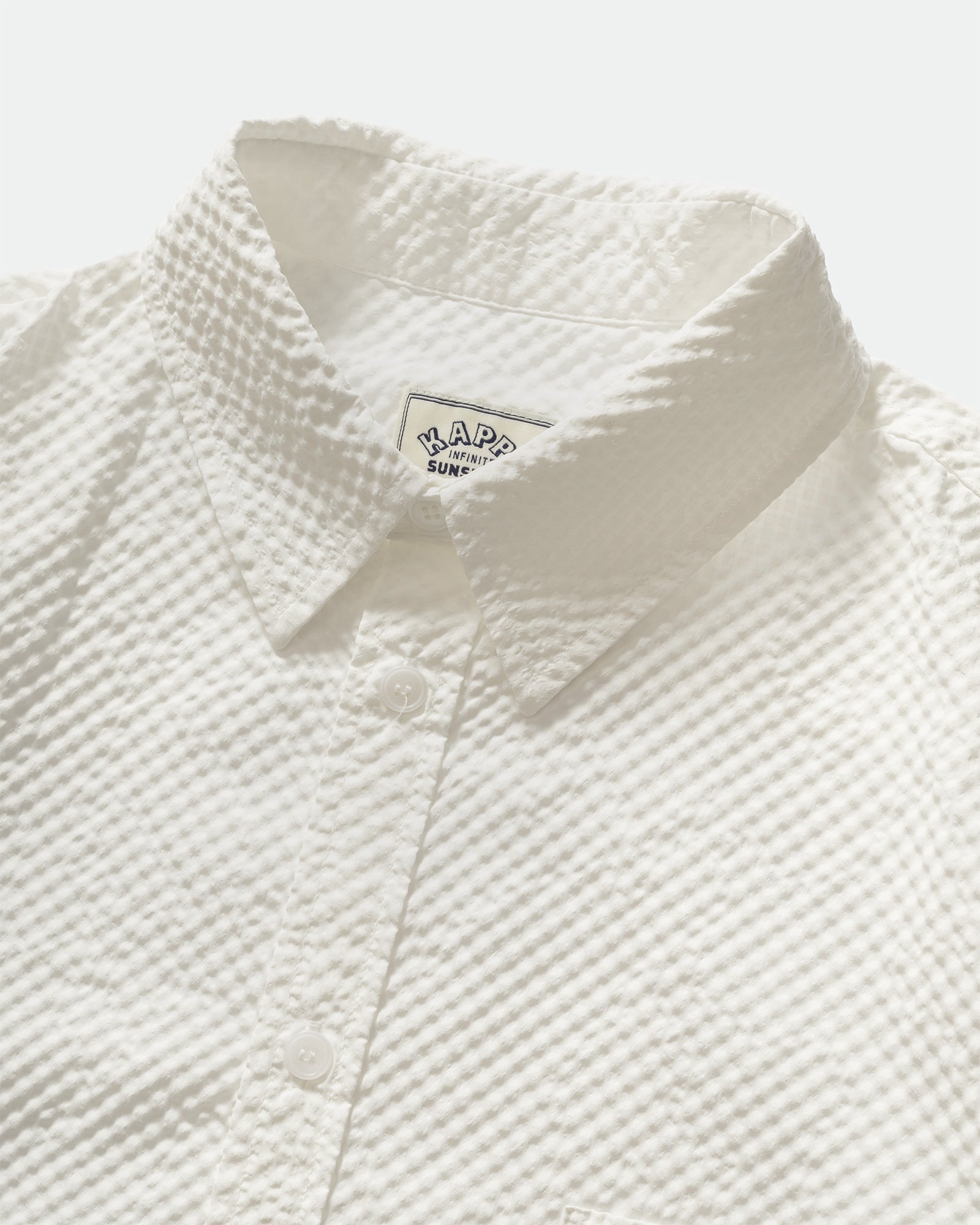 Close up of a Kappy Seersucker Check Half Shirt with a collar, branded label and  button placket on a grey background. 