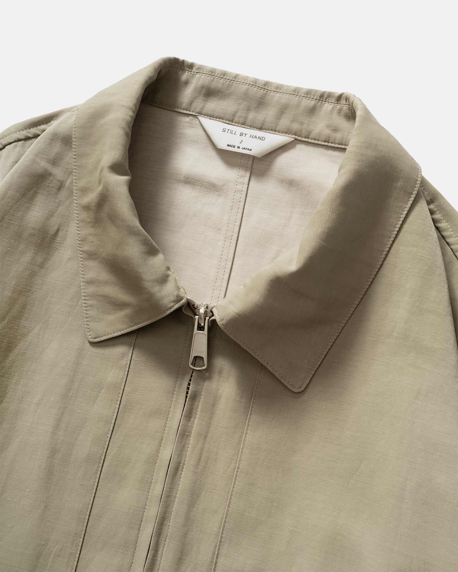Close up of Still By Hand Sage Pigment Print Zip Blouson with collar, zip closure and branded inner label on a grey background.