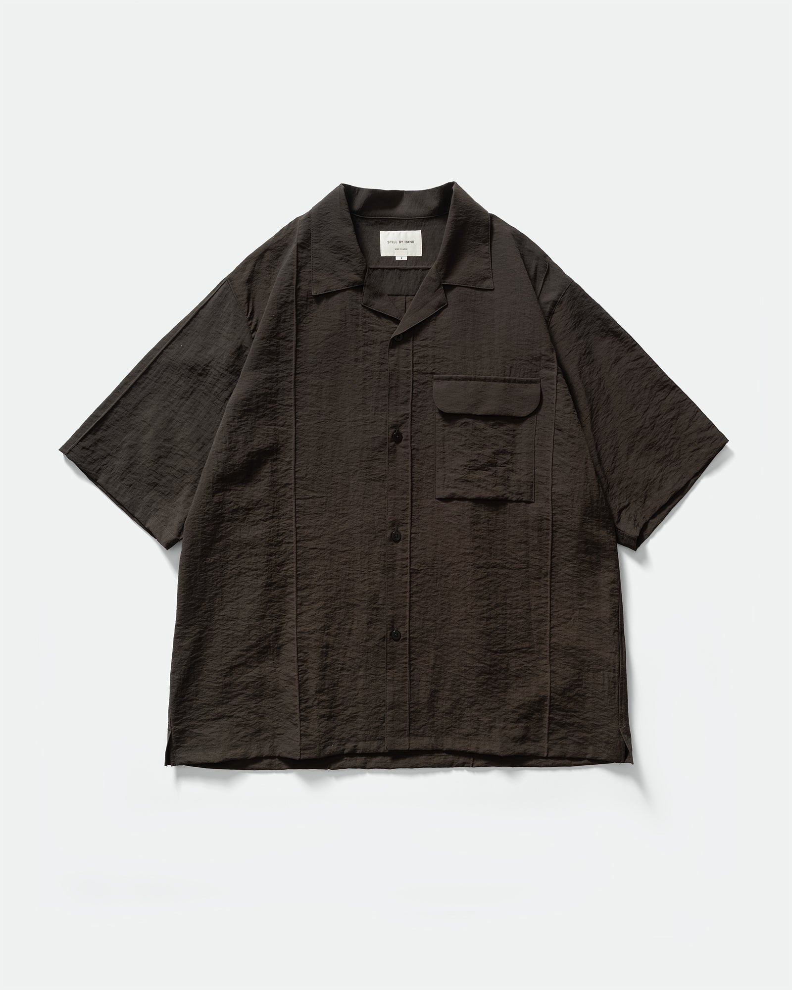 Pack shot of Still By Hand Pin Tuck Short Sleeve Shirt in Charcoal with buttons, chest pocket, collar, and branded label on a grey background.