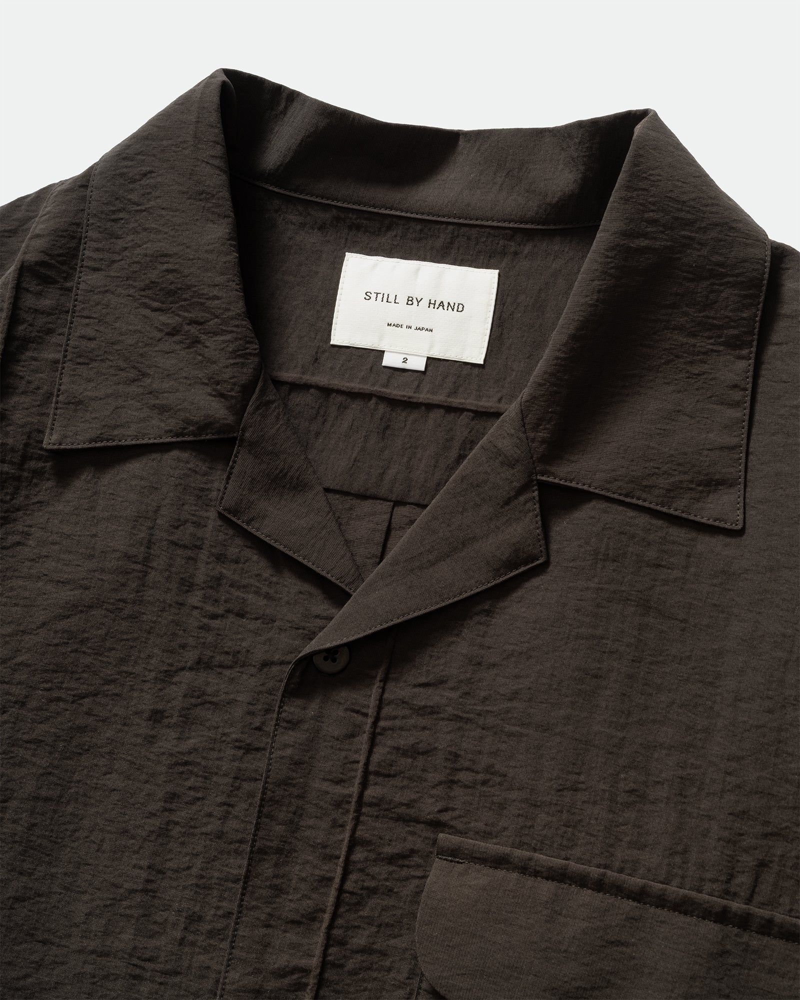 Pack shot of Still By Hand Pin Tuck Short Sleeve Shirt in Charcoal with placket, chest pocket, collar, and branded label on a grey background.
