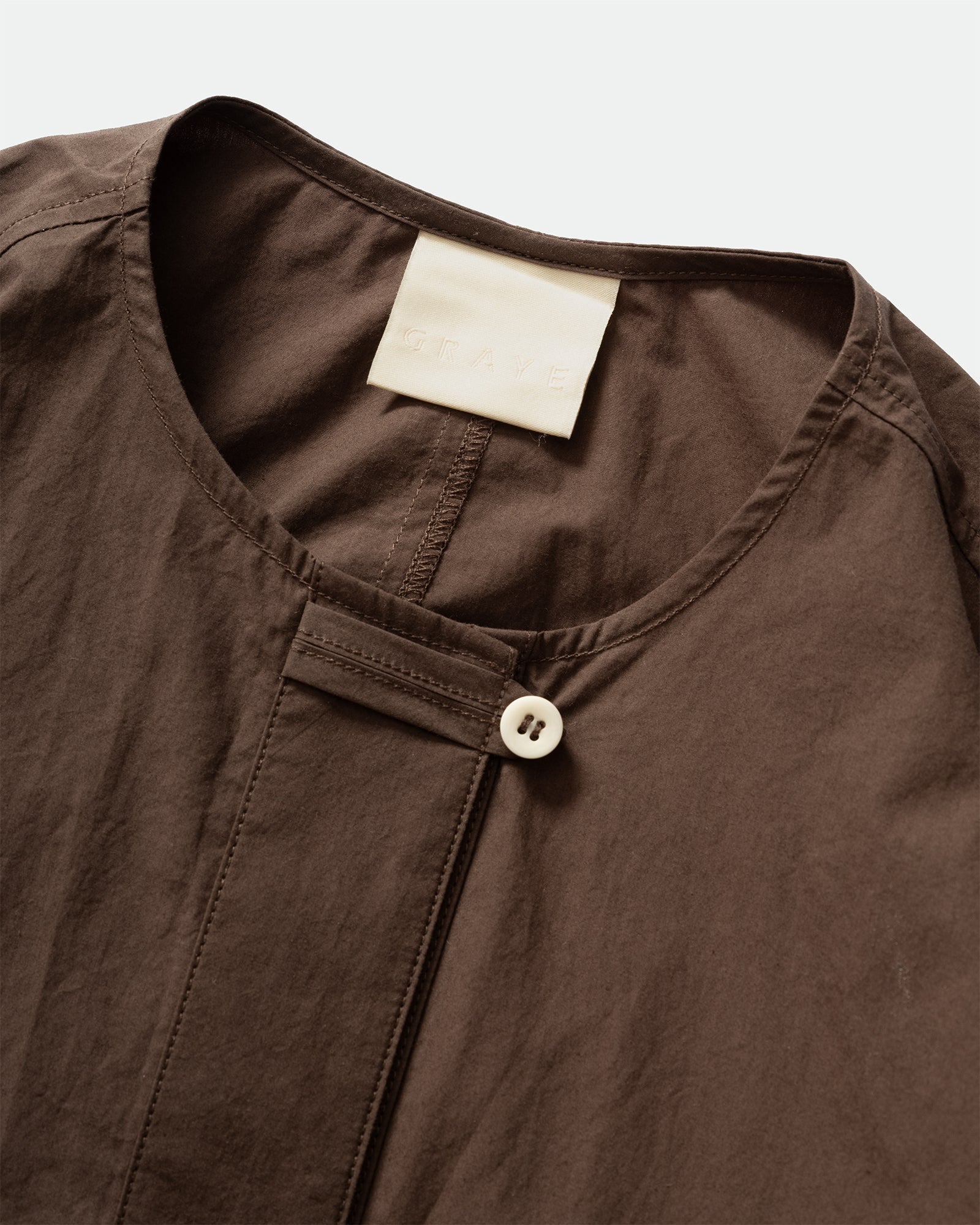 Close up packshot GRAYE Sun-Dried Cocoon Top in plum with branded label and cord loop placket on a grey background.