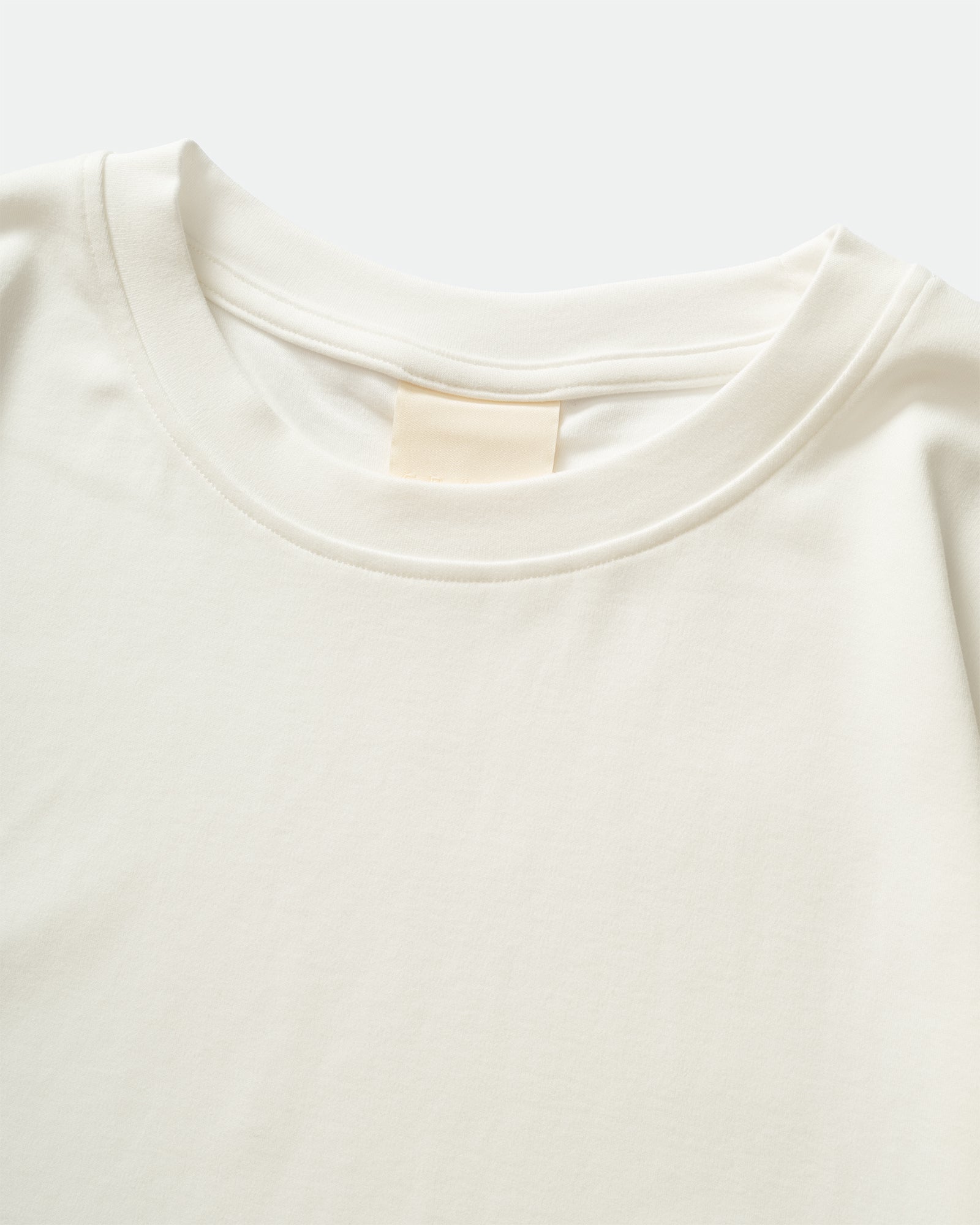 Close-up pack shot of GRAYE Cupro Signature Tee in off white with a crewneck and branded label on a grey background.