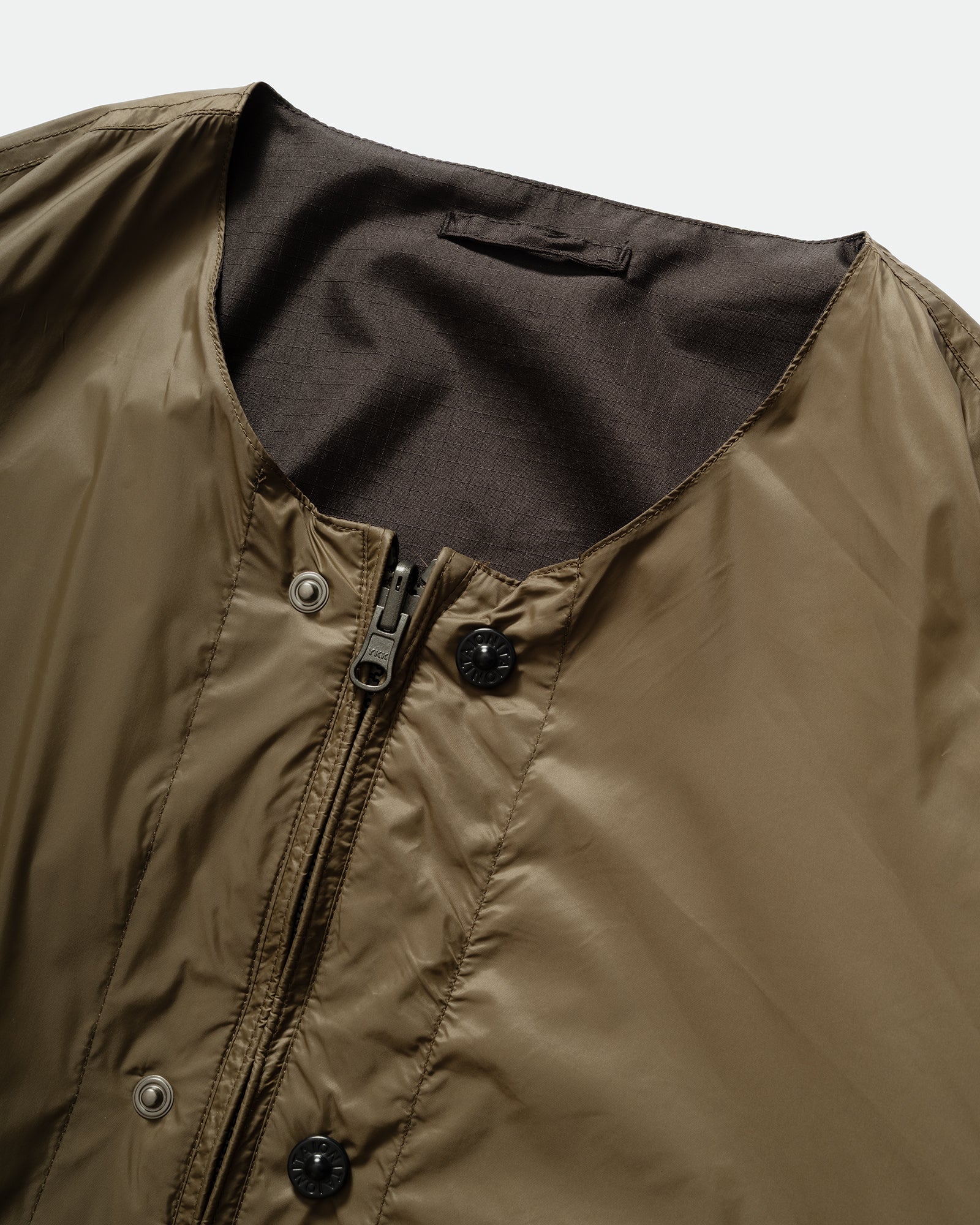 Close-up of TAION Nylon Military Reversible Liner Jacket in Dark Olive and Brown with zip closure, metal buttons crewneck on a grey background.