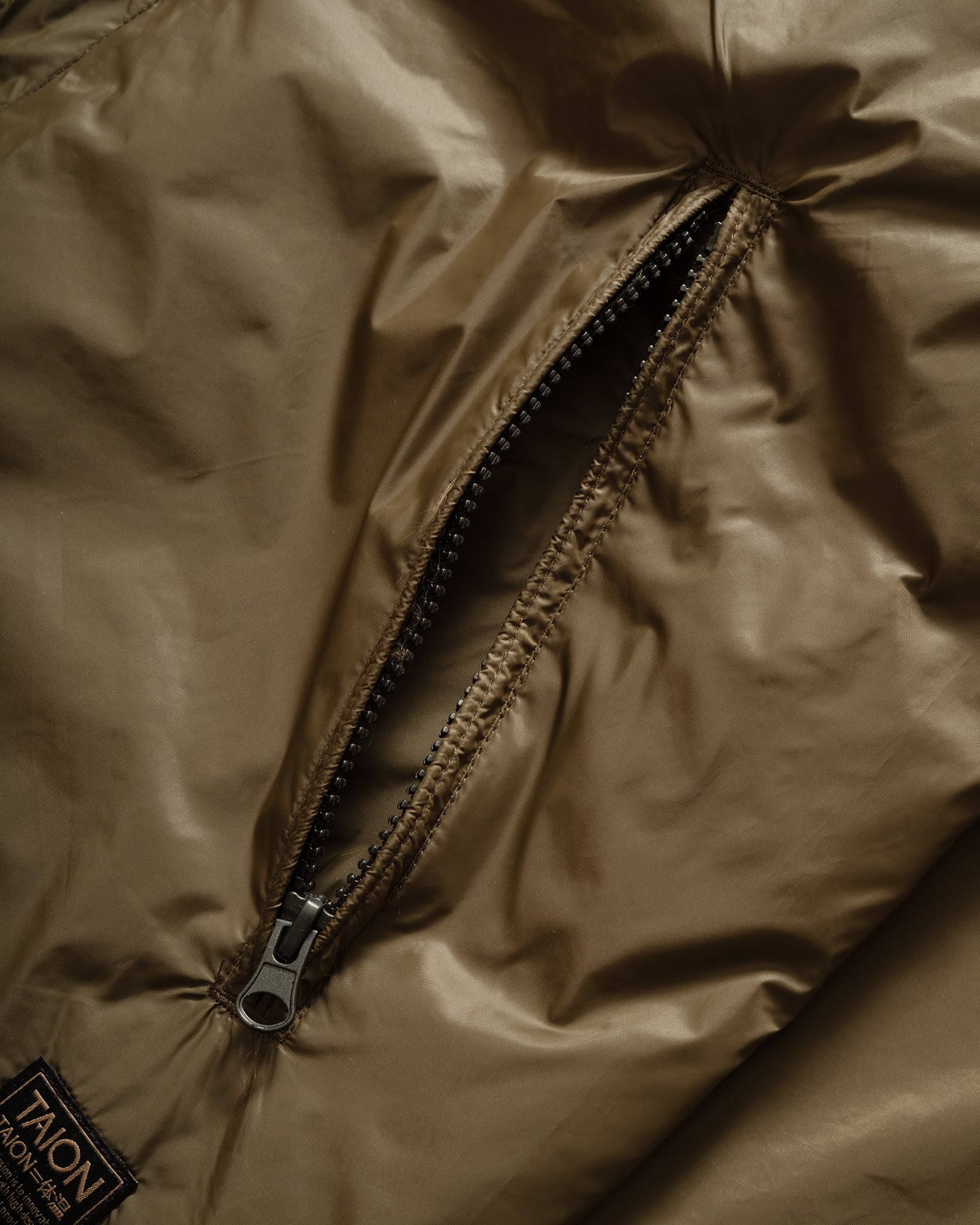 Close-up of TAION Nylon Military Reversible Liner Jacket in Dark Olive and Brown with zip pocket and metal branded embroidery.