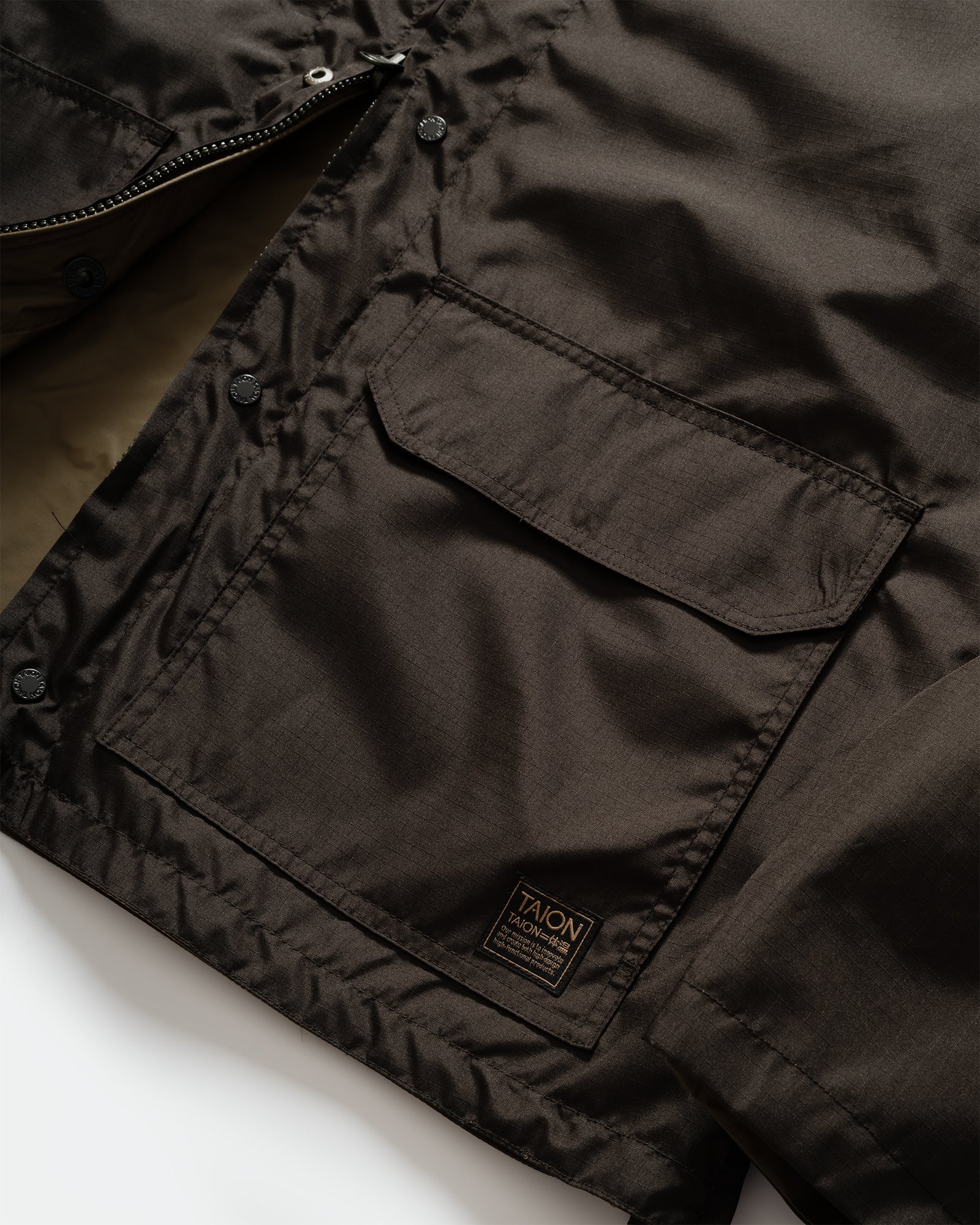 Close-up of TAION Nylon Military Reversible Liner Jacket in Dark Olive and Brown with zip closure, patch pocket, branded embroidery and metal branded buttons.